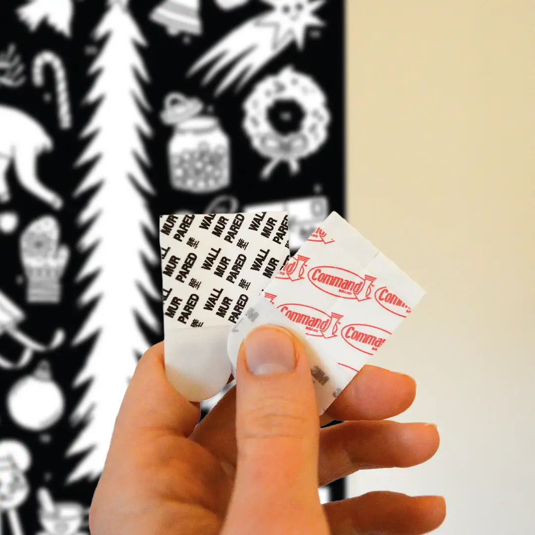Mano sujetando 4 adhesivos removibles Command para pegar el Calendario de Adviento Cocolitos a la pared (el cual se ve desenfocado en el fondo)