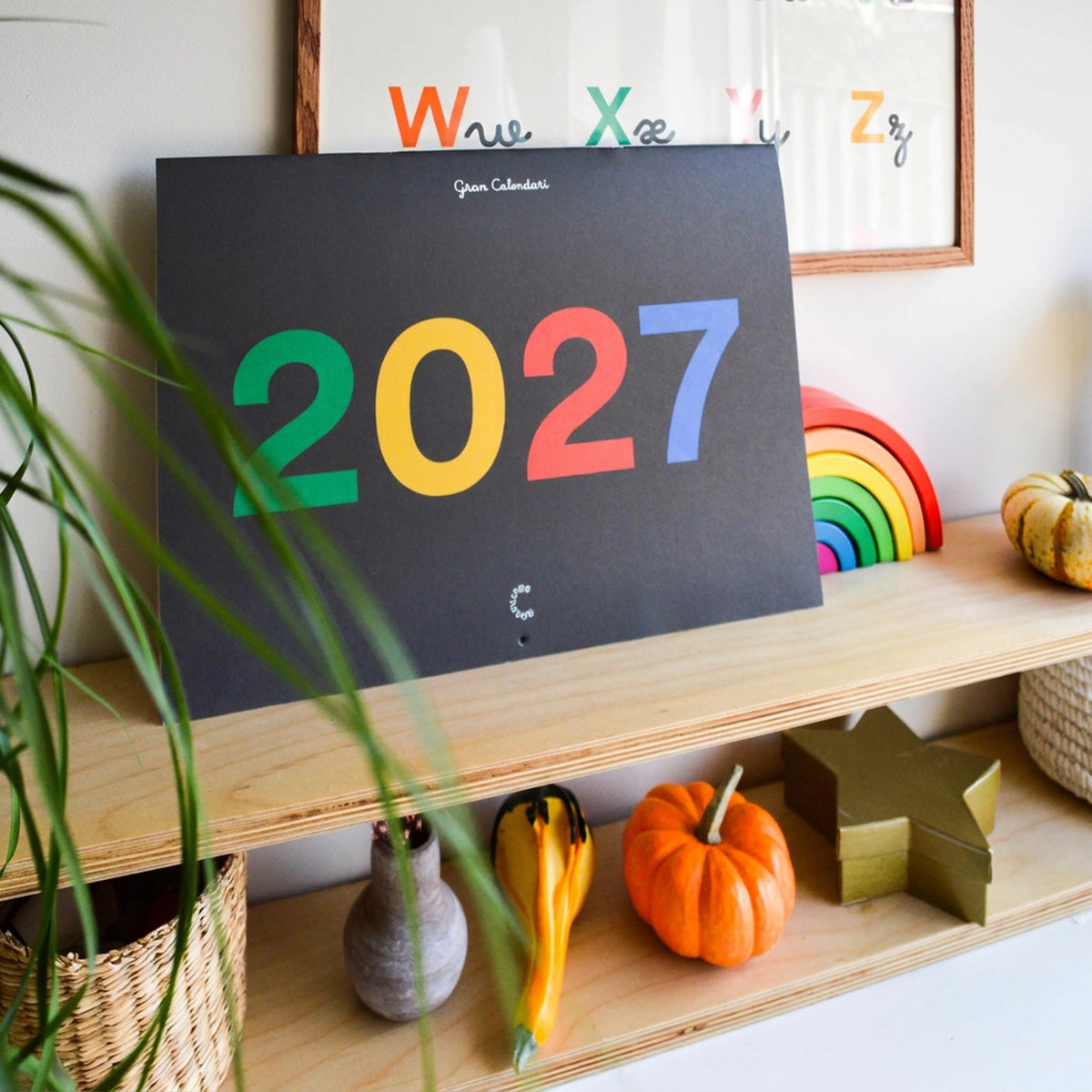 El Gran Calendari 2027 tancat, mostrant la portada, ambientat en un racó infantil, on hi apareix el pòster de l'alfabet de fons i joguines al voltant.